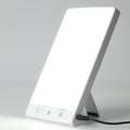 De Fashiongirl Light Therapy Lamp 10.000 lux LED is een rechthoekige lamp met een helder wit scherm, uitklapbare steun, drie bedieningsknoppen en een netsnoer aan de onderkant, ontworpen om kunstmatig daglicht te simuleren.