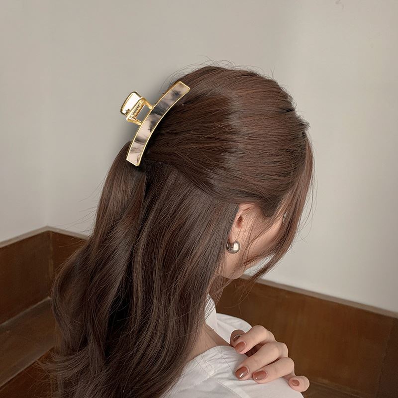 A woman with long, wavy brown hair secures her hair with the SOHO Abby Hair Claw - Ginger, pulling back the top section. She pairs it with pearl earrings and a white top—an everyday look from behind in soft lighting by SOHO.