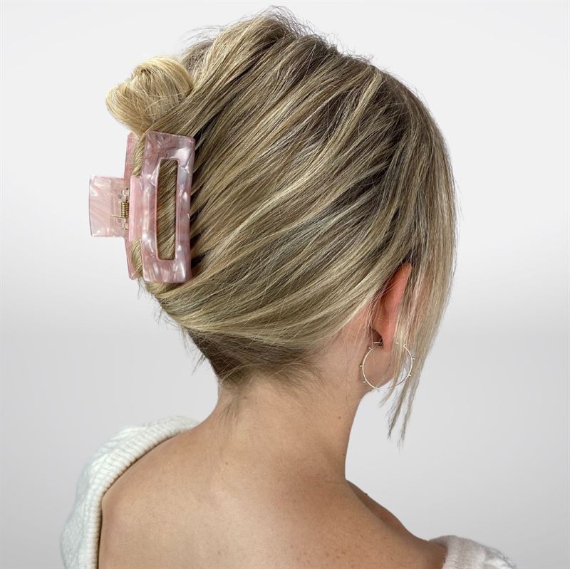 A woman with blonde hair in a low twist, secured by the SOHO Aria Hair Claw - Blush, faces away from the camera. She wears hoop earrings and a white sweater against a simple background.