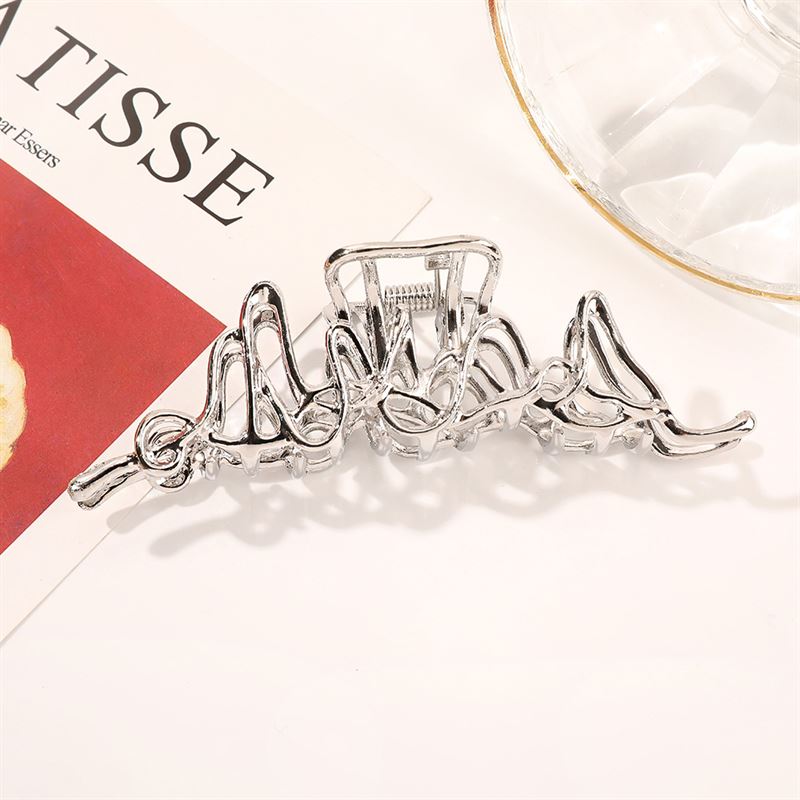 The SOHO Asli Metal Hair Claw in silver, by SOHO, features a modern wavy design and shines stylishly next to a glass dish and a magazine with "MATISSE" visible—making this accessory both chic and contemporary.