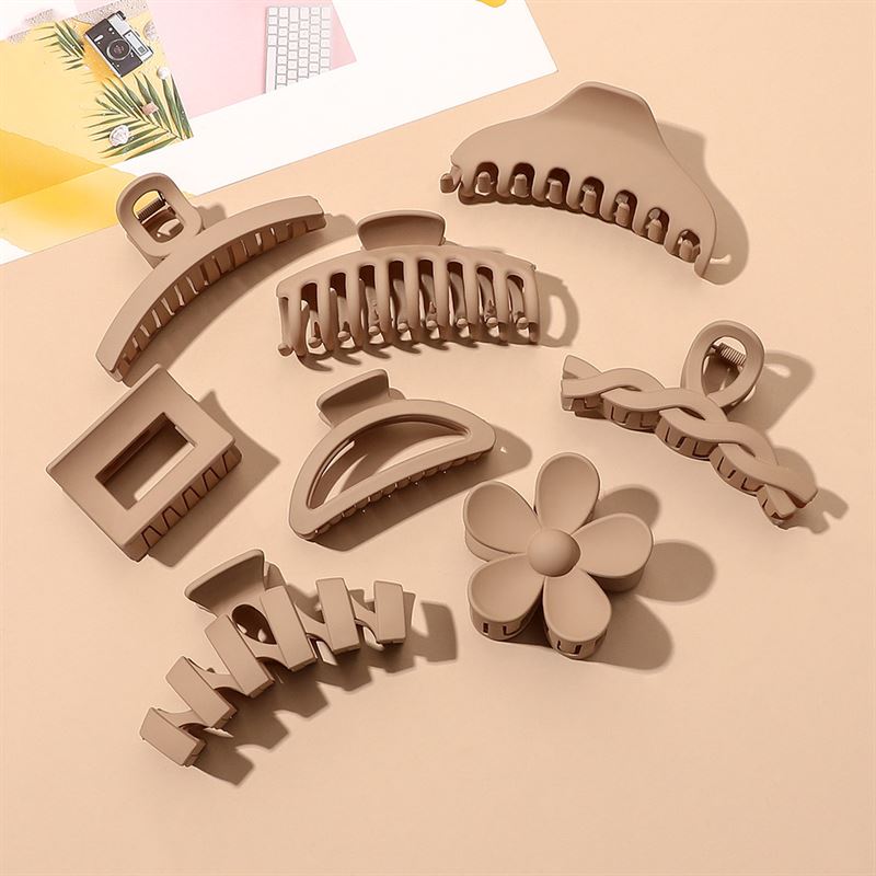 The SOHO Bloom Hair Claw - Brown by SOHO is shown with other beige and brown hair claw clips in floral and geometric shapes, arranged on a light surface with paper and a keyboard in the background.