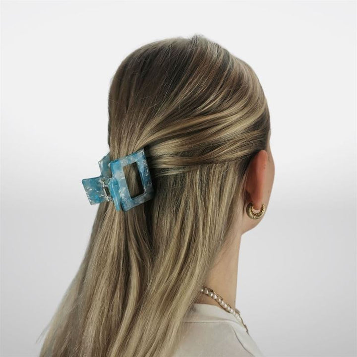 A person with long, blonde hair is seen from behind, the top section secured with the SOHO Ema Hair Claw in Crystal Blue. Gold hoop earrings and a beaded necklace complete their stylish look, showcasing this chic SOHO accessory.
