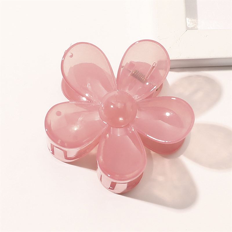 The SOHO Fiore Hair Claw - Charm Pink, a flower-shaped hair accessory with six translucent petals by SOHO, rests on a white surface near the corner of a white rectangular object.