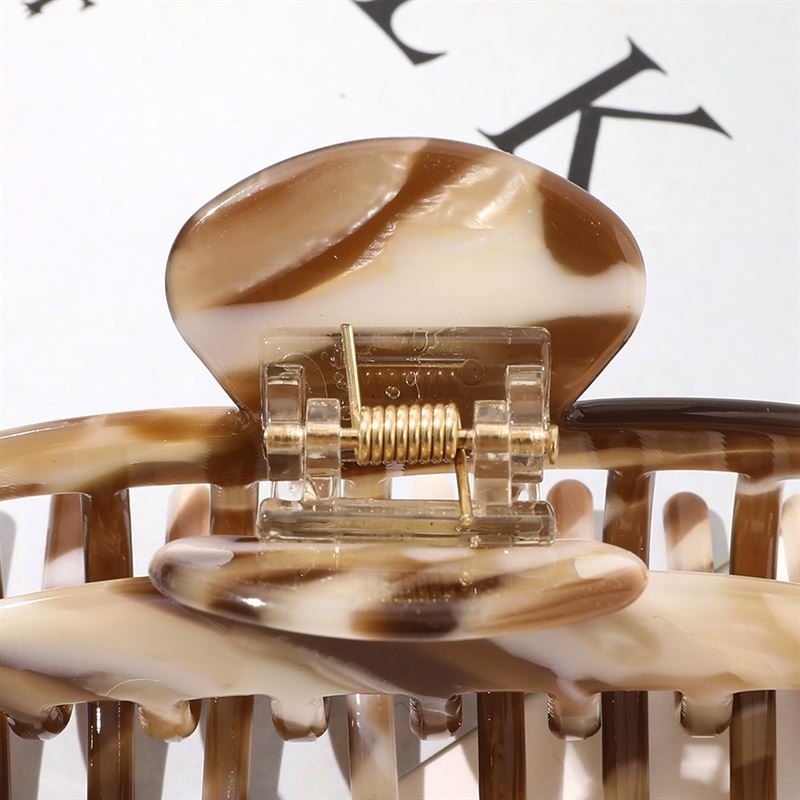 A close-up of the SOHO Gaja Hair Claw in Beige Marble—a marbled brown and cream beige hair clip by SOHO with a visible metal spring—set against a blurred background with partially visible black text.