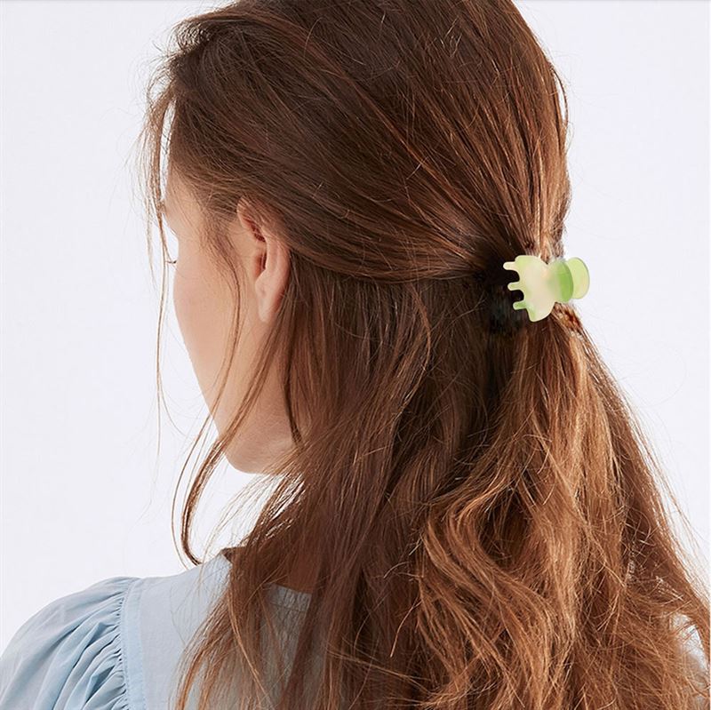 A woman with long, wavy brown hair is seen from behind, wearing a light blue blouse and securing part of her hair with the SOHO Hara Mini Hair Claw in Blush by SOHO for a secure hold.
