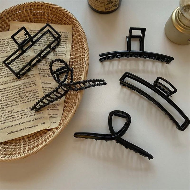 Five SOHO Iva Metal Hair Claws in black by SOHO are displayed on a white surface with a wicker tray, torn book pages, gold candle, and brown bottle—perfect for securing a messy bun or adding style like metal hair clips.