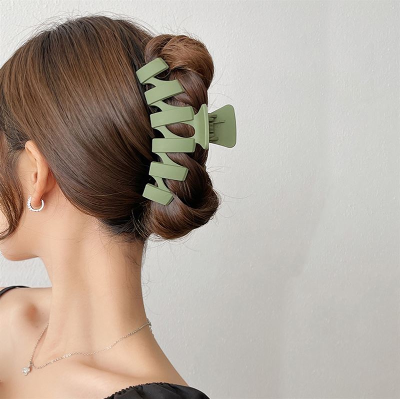 A woman with brown hair in a twisted updo, secured with the SOHO Just Hair Claw in Green, wears silver hoop earrings, a delicate necklace, and a black top against a plain light background. Brand: SOHO.