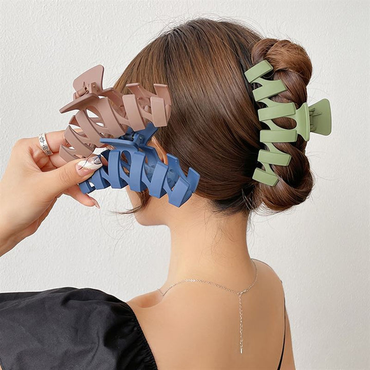 A woman with brown hair in a twisted updo, secured by the SOHO Just Hair Claw - Green from SOHO, holds two large hair clips—one brown and one blue—in her hand. She wears a black top and a delicate necklace.