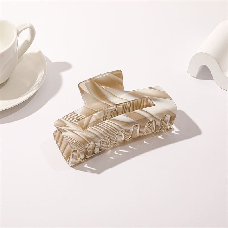 The SOHO Kaisa Hair Claw - Brown by SOHO, a marbled acetate rectangle hair accessory, rests on a white surface near a teacup and saucer, with soft shadows created by natural light.