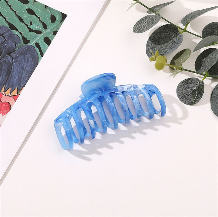 The SOHO Kana Marble Hair Claw - Ocean rests on a white surface near green leaves and a colorful painting, offering a stylish and reliable accessory.