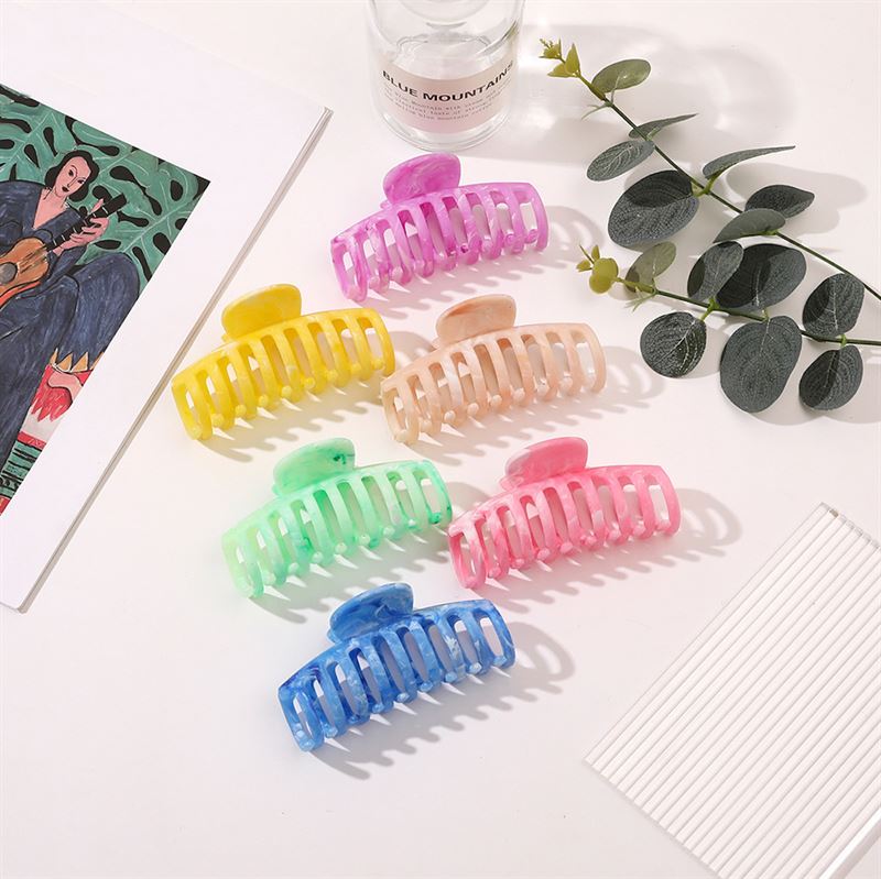The SOHO Kana Marble Hair Claw - Ocean by SOHO is displayed on a white surface. Its chic marbled design stands out among creative accents like a green leafy stem, glass bottle, striped paper, and guitar art.