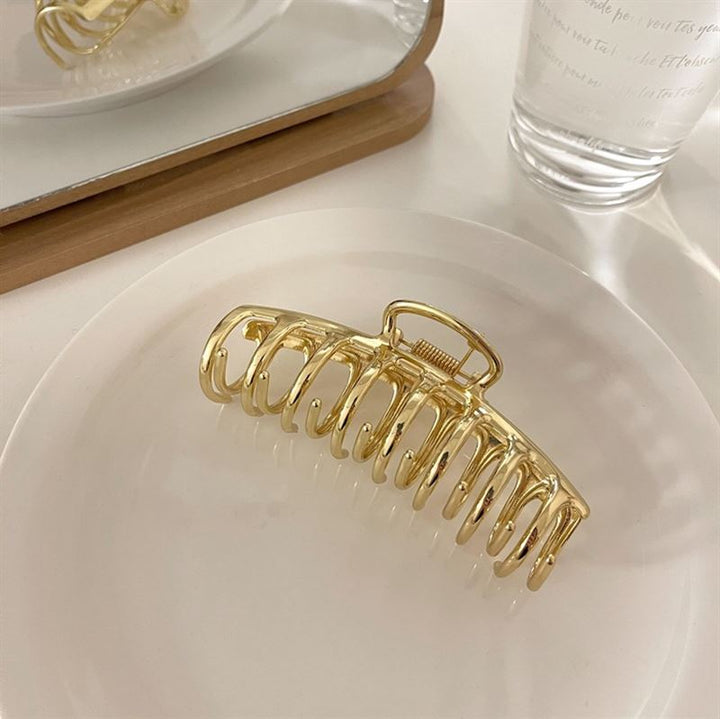 The SOHO Kana Metal Hair Claw in Gold by SOHO, designed for all hair types, features a strong grip and sits on a white plate with a mirror and clear glass of water in the background on a light surface.