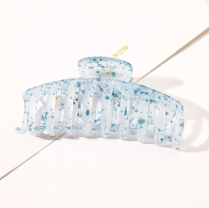 The SOHO Kana Splash Hair Claw - Blue by SOHO, featuring a translucent blue and white marbled design with a speckled pattern, offers a strong grip for all hair types. It rests on a white surface with "NOW" partially visible in the background.