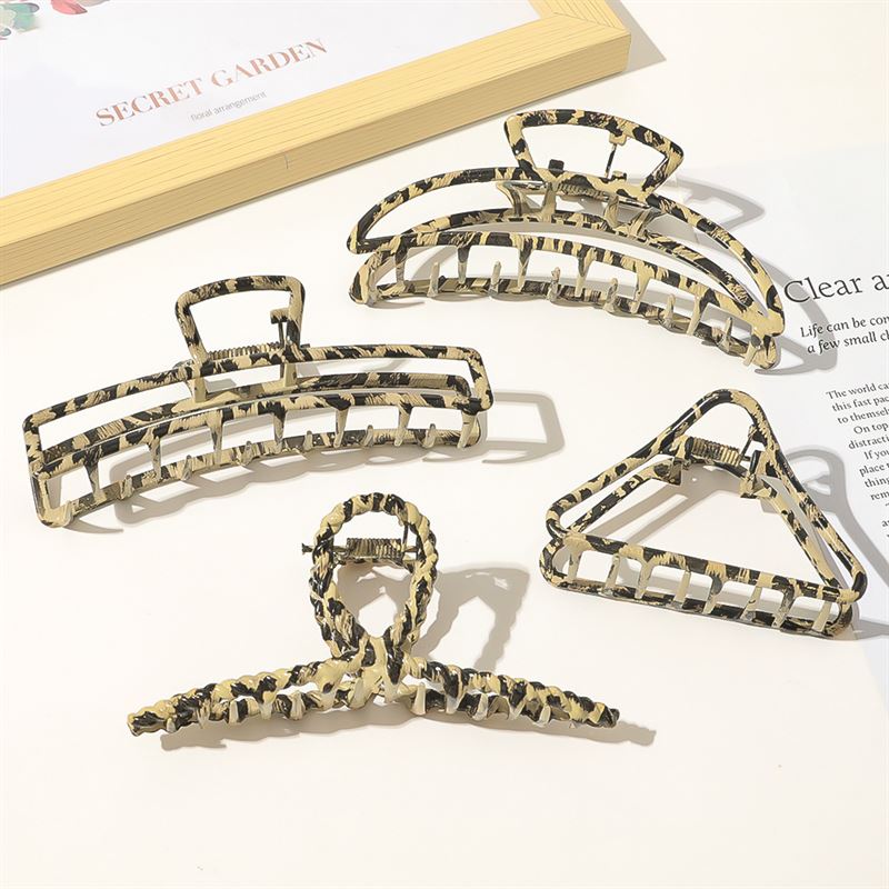 Four SOHO Leopard Metal Barette Hair Claws in gold and black geometric shapes—rectangle, triangle, oval, and bow—are stylishly arranged on a white surface near open books, showcasing chic metal hair accessories by SOHO.