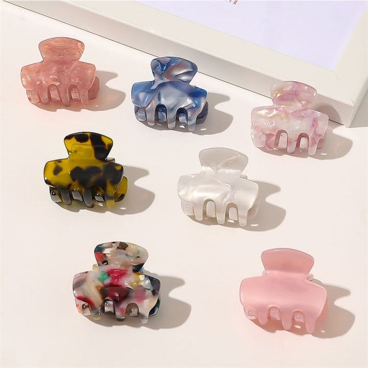 Seven colorful hair claw clips, including the SOHO Loa Mini Hair Claw in Blush, are arranged neatly on a light surface near the corner of a white-framed object.