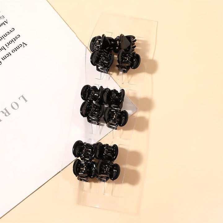 A transparent plastic pack of eight SOHO Lucca Mini Hair Claws - Black by SOHO sits on a beige surface beside a partially visible white printed sheet.
