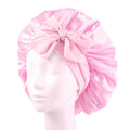 De SOHO Makena Satin Bonnet - Pink van Fashiongirl, getoond op een mannequinhoofd, heeft een glanzende roze satijnen afwerking en een grote strik aan de voorkant - ideaal voor haarbescherming en het voorkomen van kroeshaar.