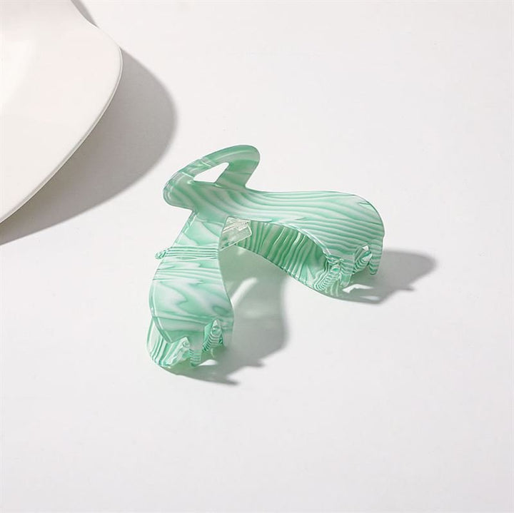 The SOHO Malou Hair Claw - Green by SOHO rests open on a white surface, its marbled green and white design casting a soft shadow. A white object appears in the top left corner.