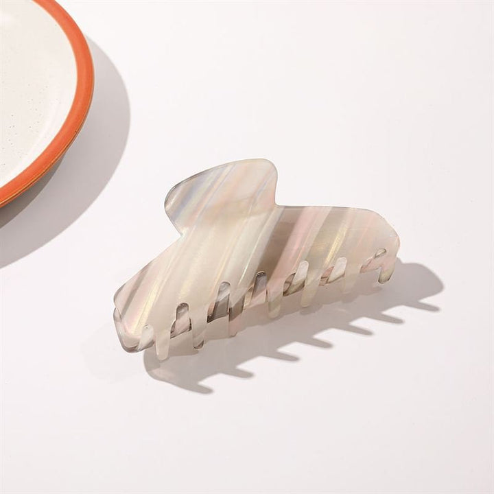 The SOHO Mesa Hair Claw - Grey Rainbow by SOHO, featuring a marbled pearlescent design, sits on a white surface near the edge of a white plate with an orange rim.