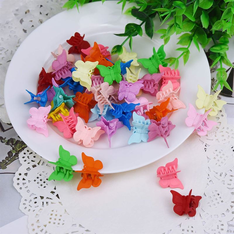 A white plate holds SOHO Mini Butterfly Hair Claws—50 multicolour hair decorations—on a lace-patterned tablecloth, with green leaves in the background. A few vibrant SOHO clips are scattered nearby.