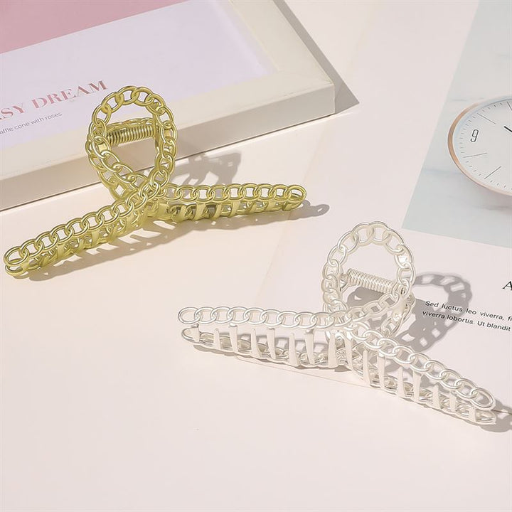 Two SOHO Monz Metal Hair Claws in gold and silver are displayed on a light surface beside a framed photo and an open magazine featuring a watch.