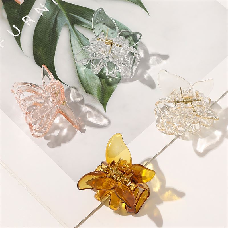 The SOHO Naja Hair Claws in clear, light pink, pale yellow, and Espresso by SOHO are arranged on a white surface with a leafy background—a stylish hair accessory for any look.