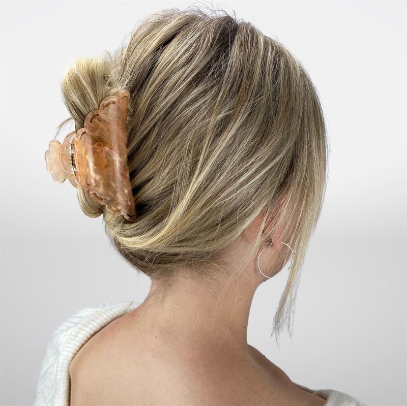 A woman with blonde hair in a low twist, secured by the SOHO Naomi Hair Claw - Sunshine, is shown from the back. She wears a white top and a silver hoop earring.