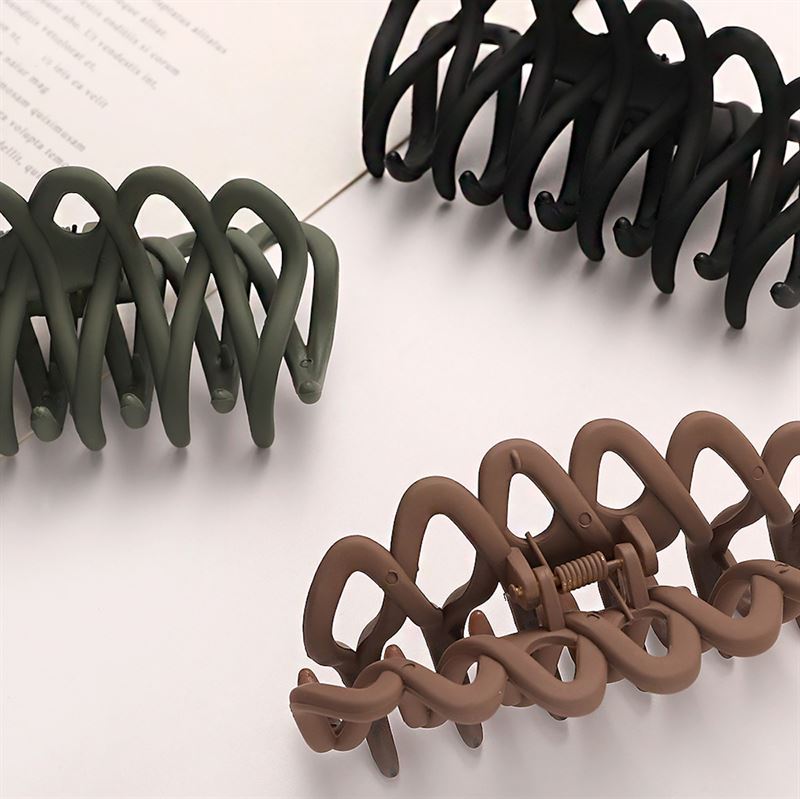 The SOHO Olive Hair Claw - Black by SOHO, a large wavy hair accessory with a gentle grip for all hair types, rests on a matte white surface with modern geometric design, partially overlapping paper with faint text.