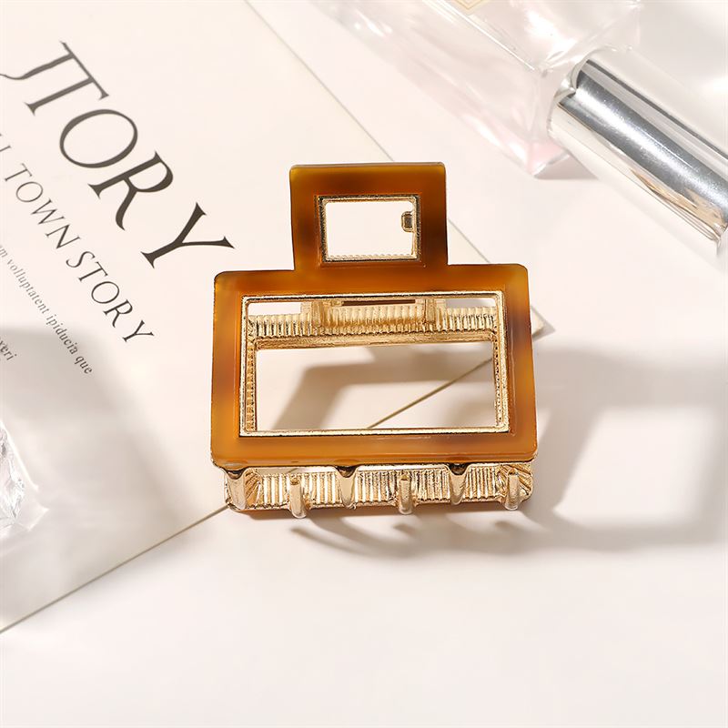 The SOHO Vida Hair Claw - Brown by SOHO, a rectangular gold and brown metal accessory with geometric cut-outs, is displayed on an open magazine with partial text and a perfume bottle nearby—an elegant accent for any look.