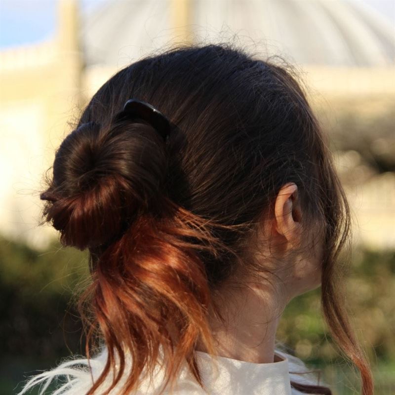 A person with brown hair in a loose bun, secured with SOHO Twist Chignon Hairpins pack of 3 and a black hair clip, is seen from behind outdoors, sunlight highlighting their hair.