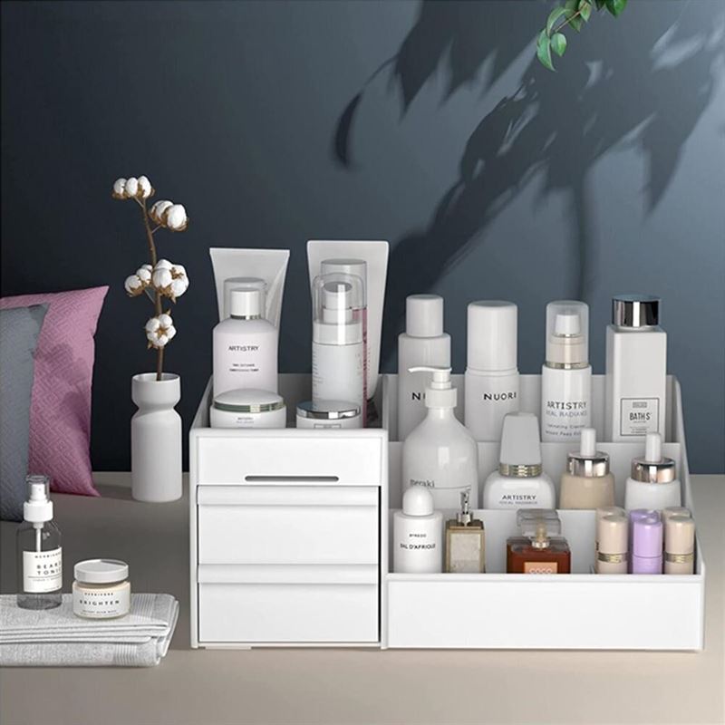 A vanity featuring the UNIQ Cosmetic Organiser with Two Drawers, White - P110 holds skincare and beauty products in bottles, jars, and tubes. The organiser keeps everything tidy, while a plant and vases decorate the background.