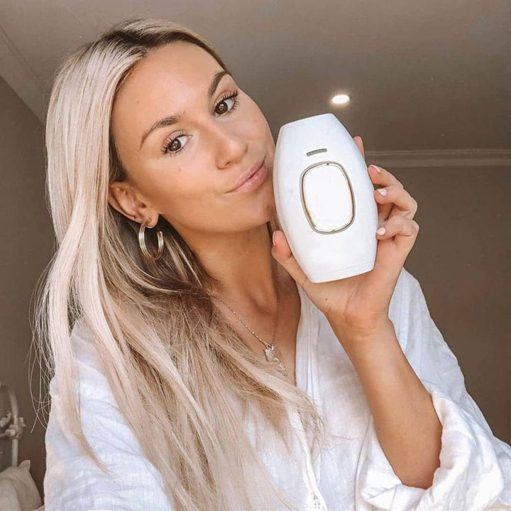 A woman with long blonde hair and hoop earrings smiles at the camera while holding the UNIQ IPL Hair Removal Device—offering smooth, pain-free permanent hair reduction at home with 600,000 flashes. She wears a white top in a bright room.