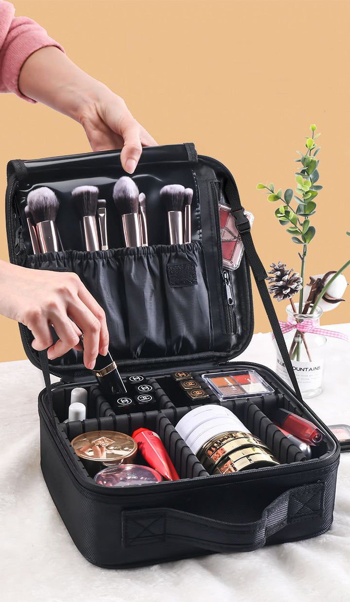 Someone organizes makeup brushes in the UNIQ makeup travel bag—an adjustable black cosmetic bag by UNIQ—filled with cosmetics, on a table beside a small potted plant.