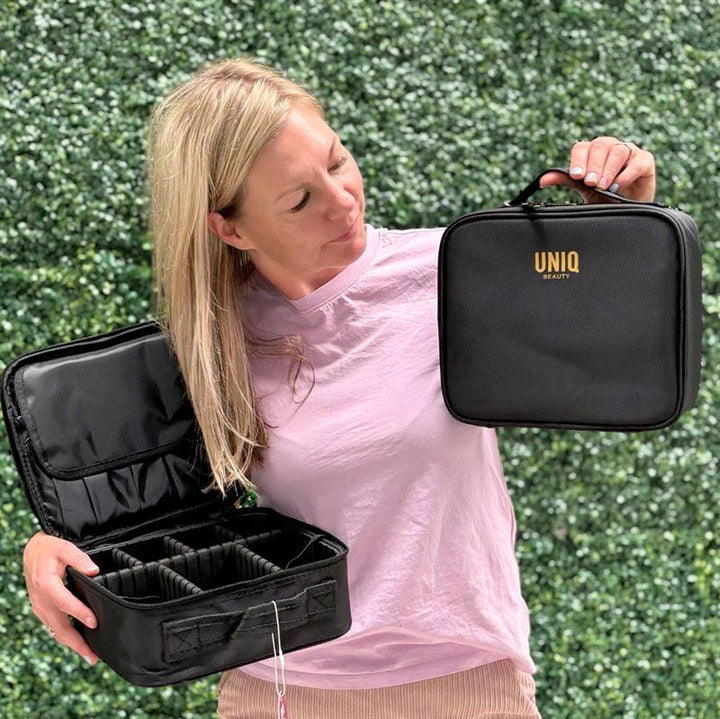 A woman in a light pink shirt holds an open UNIQ makeup travel bag with adjustable compartments in one hand and displays a closed black cosmetic bag labeled UNIQ in the other, standing before lush green foliage.