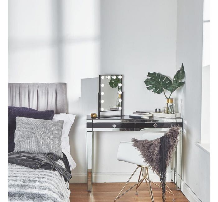 A modern bedroom corner showcases the UNIQ New York Mega Makeup Mirror with 12 bulbs in black, a mirrored vanity table, decorative plants, books, and a white fur chair beside a bed with gray and white bedding against a white wall.