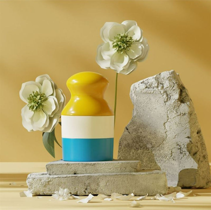 A colorful vase with yellow, white, and blue sections holds two white flowers on stacked concrete slabs beside the UNIQ Portable Refillable Sponge Roller Applicator (100ml)—a mess-free sunscreen & lotion tool for kids—set against a beige background with scattered petals.